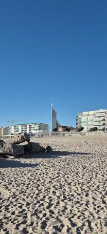 The video shows a beach scene under a clear blue sky. In the foreground, sand covers the ground, with a cluster of large rocks to the left. In the middle ground, there is a line of people walking on a paved area behind the sand. Beyond the people, there are several buildings. The most prominent building is a tall, modern church with a cross on top, situated between two apartment buildings. One apartment building on the left appears to be under construction, with a crane visible above it. The overall environment suggests a coastal town or city.