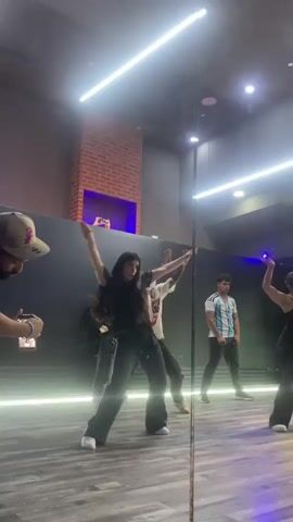 The video shows a group of people dancing in a studio. A man wearing a tan baseball cap is filming the dancers with his phone. In the center of the frame, a woman in black activewear is performing a dance move, with her arms extended. Behind her, another dancer in a black hoodie is partially visible. To the right, a man wearing an Argentinian soccer jersey is also dancing. The studio has a dark wall with a brick accent and neon lighting. A large mirror reflects the scene. The floor is made of wood.