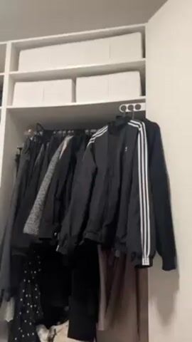A white closet is shown, with two shelves on top holding three white boxes. Below the shelves is a hanging rod filled with clothes. A black Adidas jacket with three white stripes hangs on the right end of the rod. Other clothing items such as a gray jacket, polka dot dress, and black and brown pants are visible.