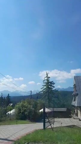 The video shows a view from a moving vehicle looking out over a rural landscape on a clear, sunny day. In the foreground, a wooden structure and a utility pole are visible. The middle ground features several white geodesic dome-like structures, trees, and some buildings. In the background, a range of mountains stretches across the horizon under a bright blue sky with scattered white clouds. The camera pans slightly, offering a dynamic view of the scenery.