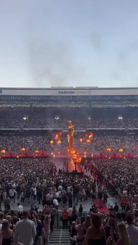 The video shows a concert taking place in a stadium. A large golden statue of a person stands on a stage in the center of the field, surrounded by flames and pyrotechnics. Thousands of people are packed into the stadium, watching the performance. The crowd appears to be enthusiastic and engaged with the show. The stadium is large and modern, with seating visible in the background. The overall atmosphere is one of excitement and celebration.
