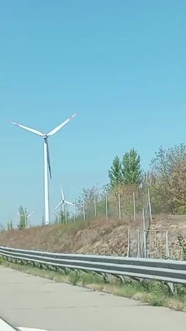 The video pans across a landscape featuring multiple wind turbines against a clear blue sky. The turbines are tall, white structures with three blades, each tipped with red. They are positioned on a slightly elevated, dry, grassy hillside. A metal fence runs along the base of the hill. In the foreground, a paved surface, possibly a road or highway shoulder, is visible with a white lane marking. Some sparse green vegetation and trees are present on the hillside, and the overall environment appears to be rural and open. The camera movement is smooth, suggesting it might be mounted on a moving vehicle.