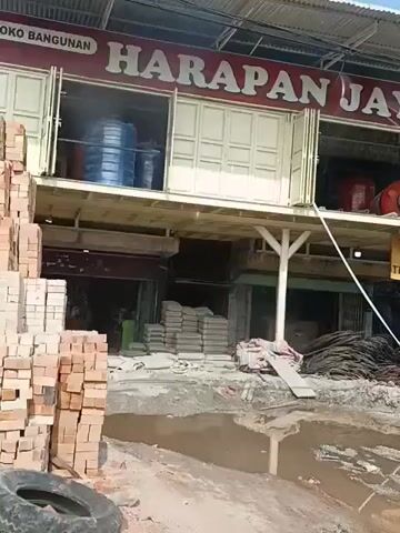 The video pans across a street scene in front of a building with the sign "HARAPAN JAYA" at the top. The building appears to be a construction supply store, with various items visible, including water tanks, red industrial machinery, piles of bricks, stacks of cement bags, and bundles of rebar. A yellow sign with black text reads "TERSEDIA DI SINI". In front of the store, there is a muddy area with standing water. The ground is uneven and appears to be a construction site or undergoing renovation. To the right of the main store, there is another storefront with a sign for "FOTO PILAR FUJIFILM". The overall impression is one of a commercial area with ongoing construction or material storage, possibly affected by recent rain.