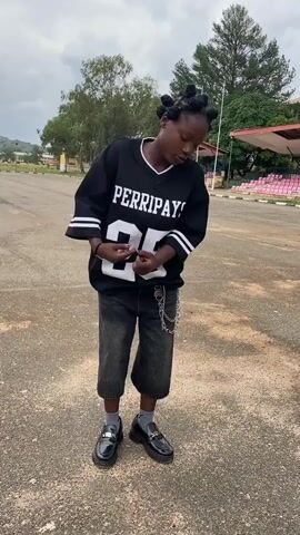 A person is performing a dance outdoors on a paved surface. The person is wearing a black t-shirt with white lettering that says "PERRIPAYS 25" and has white stripes on the sleeves, dark gray cropped pants, gray socks, and black loafers. The person's hair is styled in multiple bantu knots. A silver chain is attached to the pants. In the background, there is a stadium with pink seats, trees, and a cloudy sky. The person is facing forward and appears to be moving their arms and body.