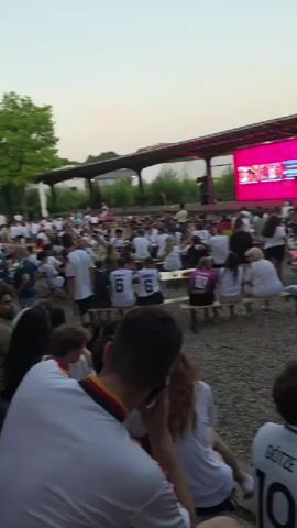 This video captures an outdoor gathering of a large group of people, likely at a public event or viewing party. The setting appears to be a park or open area with trees and a covered structure in the background, possibly a beer garden or outdoor seating area. The time of day seems to be late afternoon or early evening, with soft, natural light illuminating the scene. The weather appears to be pleasant, with no indication of rain or harsh conditions.

The subjects are predominantly people of various ages, dressed casually. Many are wearing white t-shirts, some with what appear to be national team colors (black, red, and gold) on their collars or as accents, suggesting they might be watching a sporting event, likely soccer, given the jerseys visible with numbers like "6". Some individuals are seated on benches and what look like bleachers, while others are standing. There are also tables set up, indicating a communal dining or drinking environment.

The activity taking place is clearly a social gathering focused on watching something on a large screen, visible in the background towards the right side of the frame. The screen displays colorful graphics, suggesting a broadcast of a sports match or a similar event. The overall mood appears relaxed and engaged, with people looking towards the screen. Some individuals are holding drinks, like the glass of beer visible in the foreground.

The location context of Solingen, Germany, combined with the visible jerseys and potential national colors, strongly suggests this is an event related to a German sporting team. The presence of flags (a German flag is visible on a pole in the background) further supports this interpretation. There is no significant discernible text within the video beyond the numbers on the jerseys. The focus is on the collective experience of watching an event outdoors.