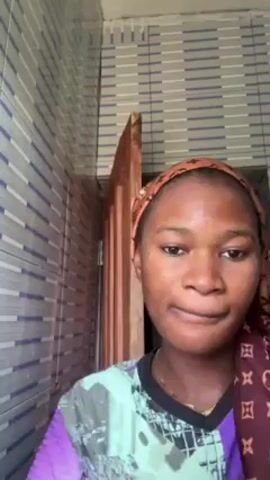 A person is applying a liquid to their face. The person is a light-skinned woman. She is wearing a multi-colored shirt and a brown and maroon head wrap. She applies a drop of liquid to the upper portion of her cheek. The liquid comes from a brown glass bottle with a black dropper. She rubs the liquid into her skin. The background includes a vertically striped tiled wall, a partly visible door, and a ceiling with horizontal bars. The person is facing the camera for the entire video.