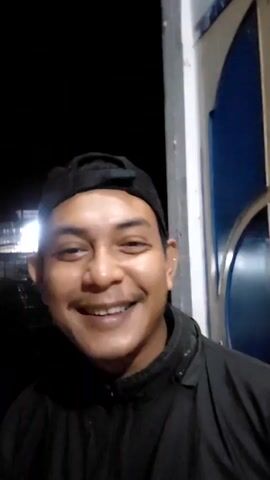 A man with brown skin and a black cap stands facing the camera. He has a mustache and is wearing a black jacket. He is smiling and talking to the camera. Behind him is a white pillar next to a door painted blue and white. The background behind the door is dark.