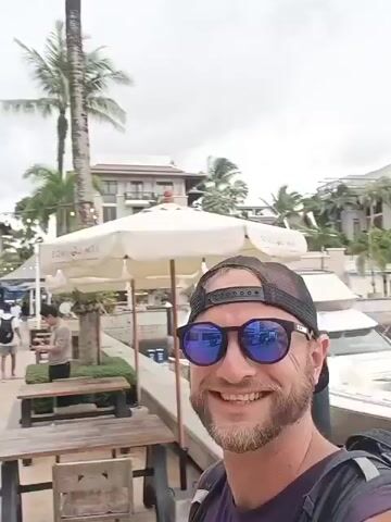 A man with a beard, sunglasses, and a baseball cap takes a selfie with a marina full of yachts and buildings in the background. The man is smiling and wearing a purple t-shirt and a backpack. The sky is overcast. The video is shot from a first-person perspective.
