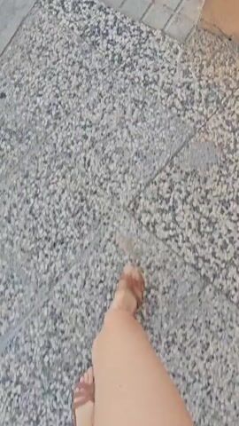 The video begins with a close-up shot of a person's bare leg and foot wearing a brown sandal, walking on a tiled sidewalk. The camera then pans down to follow the person's leg and foot as they walk along a concrete curb. The scene transitions to the person walking on an asphalt road, with the camera continuing to focus on their leg and foot. Finally, the video shows the person walking past a manhole cover on the asphalt road. The lighting suggests it is daytime.