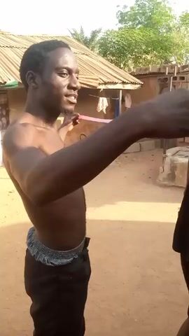 A shirtless, dark-skinned man is standing outdoors in front of a person who is mostly out of frame. The man holds a pink measuring tape between his outstretched hands. The tape hangs vertically and is marked with numbers in a light-colored ink. The man is looking at the measuring tape and speaking. Behind him is a sandy ground, a building with a corrugated metal roof, and a pile of bricks. A few trees are visible in the background. The overall lighting appears to be sunny and bright.