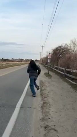 A single person, viewed from behind, is running down a paved road. They are wearing a dark jacket and blue jeans. The road has a white line marking the edge. To the right of the road is a sandy shoulder, a wooden fence, and vegetation.  Overhead are power lines supported by poles. The sky is overcast. At one point, the runner briefly leaps into the air.