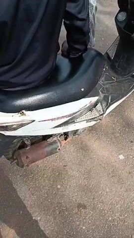 A person wearing a black jacket and sitting on a black scooter seat is visible.  The scooter is white and black with red accents, and it appears to be damaged. The scooter's exhaust pipe is rusted. Another person wearing white patterned pants stands near the scooter. The ground is asphalt.