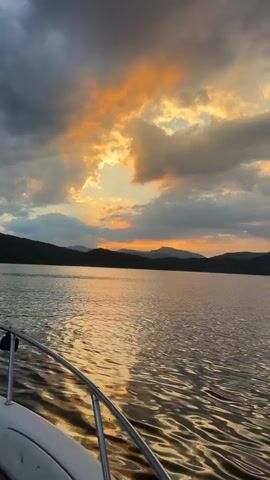 The video shows a boat moving on a lake at sunset. The sky is dramatic with dark clouds interspersed with bright orange and yellow light from the setting sun. The sun is partially obscured by clouds, creating a glowing effect. Hills or mountains are visible in the distance across the lake. The water is rippling, reflecting the colors of the sky. The bow of the boat is in the foreground, with a metal railing visible. The boat appears to be white with brown seating. The camera is positioned on the boat, looking forward.