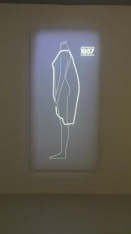 A video displays a digital projection of a stylized human figure wearing a dress outline, displayed on a vertical white canvas. The projection shows the figure from a side profile, facing left. The figure is outlined in a bright, thin line that appears to be pulsing or glowing with a light green hue. The outline depicts a headless torso with arms held in front and legs extended. The dress outline itself is a voluminous, A-line shape that extends from just below the neck area down to just above the knees. The date 1957 is illuminated in the upper right corner of the projection in a similar glowing line. The canvas is mounted on a light-colored wall, with the bottom portion of the canvas extending out of the frame. The environment appears to be an art gallery or exhibition space, with soft, ambient lighting contributing to the overall muted tones of the background. The video captures a slow, subtle movement within the projection, suggesting a dynamic display rather than a static image.