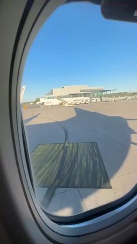 The video opens with a view from an airplane window showing a bright, sunny day at an airport. A terminal building is visible across the tarmac, which features yellow directional arrows and a large green rectangular marking with black lines. Long shadows are cast, suggesting it's either early morning or late afternoon.

The scene then shifts inside the airplane cabin, looking down an aisle where passengers are seated in yellow and dark blue seats. Overhead compartments and ceiling lights are visible. An audio announcement in German is playing, mentioning "Hinweisschildern und Anweisungen der Besatzung" (signs and instructions of the crew). A man in a black hoodie is seen looking at his phone.

The video concludes with a close-up selfie of a man wearing a black leather jacket and a black cap, smiling slightly. Another passenger is visible behind him. His jacket has some blurry white text or graphics. The overall setting is consistent with an airplane on the ground at an airport, likely in Weeze, Germany, given the location context and German announcement.