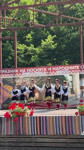 A group of six young females in traditional Bulgarian folk costumes are standing on a wooden stage, performing a dance. They are all wearing white blouses with embroidered trim and vests with colorful patterns, black skirts, and red tights. Each performer has flowers in her hair. The stage is decorated with a striped fabric banner and potted red flowers in the foreground. Behind the performers, a banner with Cyrillic text is displayed against a backdrop that depicts a stone wall and a building. The scene appears to be outdoors, with trees visible in the background. The overall impression is of a cultural festival or performance.