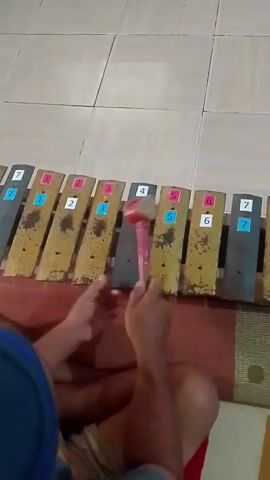 The video shows a person playing a xylophone. The xylophone has metal bars of varying lengths and colors, with numbers and colored stickers on them. The person is using a red mallet to strike the bars, producing musical notes. The person is sitting on the floor, and the xylophone is placed in front of them on a mat. The background appears to be a tiled floor. The person is wearing a blue shirt and shorts. The video is filmed from a top-down perspective.