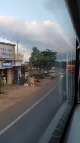 The video shows a view from a window, likely a bus, looking out onto a road. The road curves to the right and appears to be a two-lane asphalt road with a white line dividing the lanes. On the left side of the road, there are some buildings, trees, and various objects. One building has signs that appear to be advertisements. There is a small structure covered with a tarp. Several vehicles are visible on the road, including cars, trucks, and a motorcycle. The sky is partly cloudy with shades of blue and white, and the lighting suggests it is either early morning or late afternoon. The camera is moving, giving the impression of traveling.