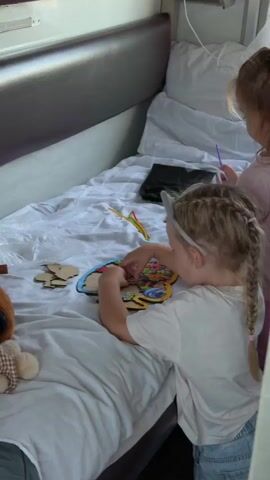 The video shows two young children playing on a bed in what appears to be a cabin or a small room. The room has a bunk bed setup with a grey upholstered headboard and white bedding. A teddy bear is visible on the left side of the bed.

The primary subject is a young girl with blonde hair in two braids, wearing a white t-shirt and denim shorts. She is actively engaged in assembling a colorful wooden puzzle depicting cartoon characters. She has a headband with what look like cat ears on. Beside her, another young girl, with blonde hair tied in a ponytail, is also looking at the puzzle and holding a purple stick. Several pipe cleaners in red, yellow, and green are scattered on the bed near the puzzle.

The activity captured is the children playing with a puzzle and other craft materials. The scene suggests a daytime setting, possibly indoors for comfort or travel. The children appear focused and engaged in their play. There is no visible text in the video. The setting might be a train or a boat cabin, given the enclosed space and the way the bed is built into the wall.