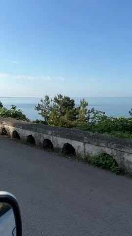 A vehicle travels along a road beside a stone wall with arched openings. The wall runs parallel to the road and the sea beyond. Trees and lush green vegetation border the road and the wall. The sky is a clear blue.