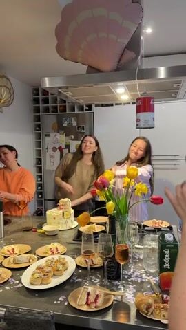 Three women are gathered around a kitchen island, which is covered with plates of cake, pastries, champagne glasses filled with rose wine, a bottle of wine, and a carton of juice. They are clapping and singing along to music. Two balloons decorate the scene, one shaped like a pink shell and another, a can of tomato soup, hangs from the ceiling. The kitchen has white cabinets and stainless steel appliances. A wall of wine bottles is built into the cabinets. The women are dressed casually, one in an orange sweater, one in a beige sweater, and one in a pink striped button-down shirt.  A vase with yellow and red tulips sits in the middle of the island. The countertop is covered with metallic confetti.