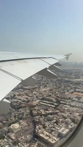 The video shows an aerial view of a city from an airplane window. The airplane's wing is visible in the foreground, angled across the frame. Below, a dense urban landscape is spread out, characterized by numerous buildings with light-colored roofs. Streets are visible winding through the city, with some appearing to have vehicle traffic. In the distance, a hazy skyline of taller buildings suggests a more modern part of the city. The sky is clear and light blue, indicating a daytime scene. The perspective is looking down and out from the aircraft.