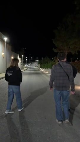 Three individuals are walking down a paved road at night. The person on the left is wearing a black hoodie and blue jeans. The person in the middle is wearing a flannel shirt, blue jeans, and a crossbody bag. The person on the right is wearing a dark jacket and blue jeans. Streetlights illuminate the road. The road appears to slope gently downhill. Cars are parked in the distance. There is a low wall lined with shrubbery to the right of the road.