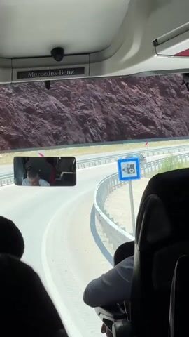 The video is filmed from inside a bus looking out the front windshield. In the rearview mirror, a person wearing a white shirt and dark pants is visible, leaning forward with their head down, seemingly asleep or resting. Outside the bus, a winding road is seen with a guardrail along the side. Several cars are driving on the road. The landscape outside appears to be mountainous with rocky cliffs. The interior of the bus shows the backs of passenger seats.