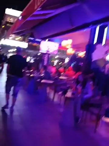 The video captures the interior of a bar or restaurant with a lively atmosphere. The lighting is predominantly blue and purple, creating a nightclub-like ambiance. There are tables and chairs arranged throughout the space, with patrons seated at some of them. Several people are visible, some appearing to be seated, while others are moving around. There are multiple television screens mounted on the walls, displaying content. The space is decorated with hanging lanterns that provide warm, orange light. The overall impression is that of a busy and vibrant social environment.