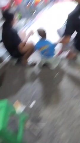 The video shows a group of people playing with toy cars on a race track. In the foreground, two people, an adult and a child, are sitting on small stools, facing the race track. The adult is wearing a black t-shirt with a graphic on the back and a green cap. The child is wearing a blue t-shirt. The race track is elevated and appears to be made of white material with red and blue borders. There are also other toy cars visible in the background. The environment appears to be outdoors, with a rough, dirt-like surface on the ground. The video is shaky and the lighting is dim, suggesting it was filmed in low light conditions.