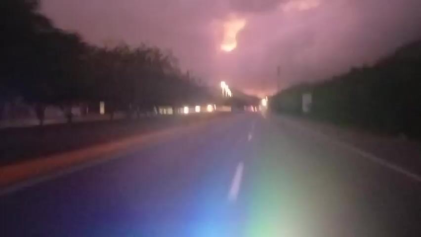 The video shows a point-of-view shot from a vehicle driving on a road at dusk. The sky is dark with purple and pink hues, and there are clouds. A faint lightning flash is visible in the sky. The road has white lane markings and a yellow line on the left side. Trees and some buildings line the side of the road. As the video progresses, lights from oncoming vehicles and streetlights become more apparent. Towards the end of the clip, the dashboard of the vehicle, with its illuminated gauges, comes into view. The overall atmosphere is that of driving in stormy weather at night.