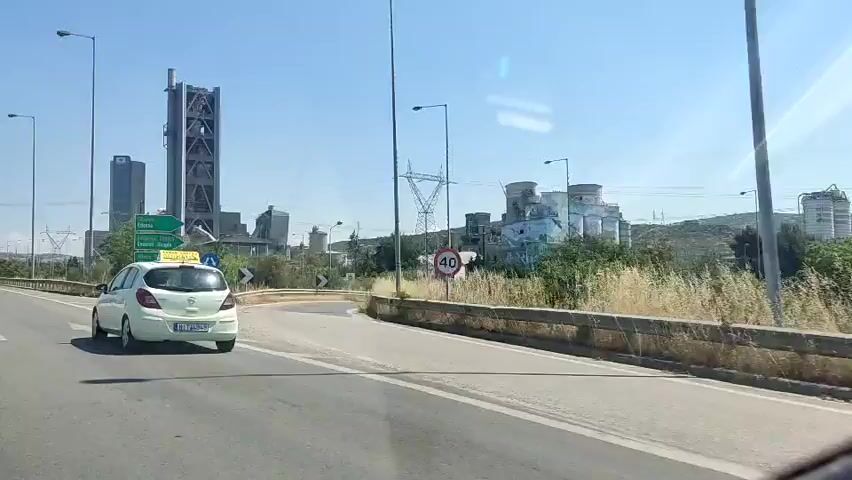 A dashcam video shows a white car driving on a highway. A car is in front of the dashcam car, and a large factory with tall structures and silos is visible in the background. The factory appears to be a cement plant. A speed limit sign with "40" is visible on the right. Green signs with white text point to various locations including "EKOZONA EDESSA," "ERZONDI SKOPIA," and "ATHENA VEROIA." The sky is clear and blue.