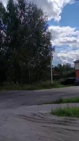 The video shows a static, side-view of an outdoor scene. The environment is rural or semi-rural, with trees and grass. There is a paved road or pathway in the foreground, and a light pole visible on the left side. The sky is cloudy with patches of blue. The perspective suggests the video might have been taken from a moving vehicle, but the scene itself is not actively changing. There are no people or significant actions depicted.