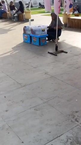 A person wearing a headscarf and dark clothing is kneeling next to two blue cooler boxes filled with bottled drinks. The cooler boxes are stacked on top of each other. A metal stand with a black base and a vertical pole is positioned in front of the person, and the person appears to be interacting with the contents of the cooler boxes. The ground is paved with light-colored square tiles. In the background, other people are seated and some structures are visible. The scene is outdoors, with sunlight casting shadows on the tiled ground.
