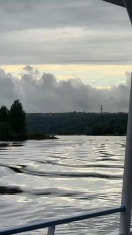 The video captures a serene view from a boat moving through calm water. The water's surface is disturbed by gentle ripples, reflecting the soft, diffused light of an overcast sky. Lush green trees line the distant shore, creating a verdant backdrop. In the far distance, a solitary structure, possibly a tower or pylon, stands against the horizon. The sky above is filled with layers of clouds, with a hint of lighter sky visible through them. The foreground is occupied by the edge of the boat, with a white railing and blue trim. The overall impression is one of peaceful transit through a natural landscape.