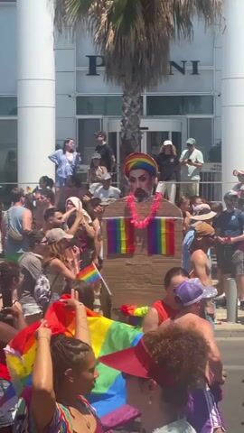 A crowd of people are gathered outdoors, many holding or waving rainbow flags, indicating a Pride event. In the foreground, a large cardboard cutout of a man wearing a rainbow headdress and a lei is being carried. Behind it, a double-decker bus with "GENDER BLENDER" written on its side is visible, with people on top. The event appears to be taking place on a sunny day in a city with modern buildings. Some individuals are filming with their phones.