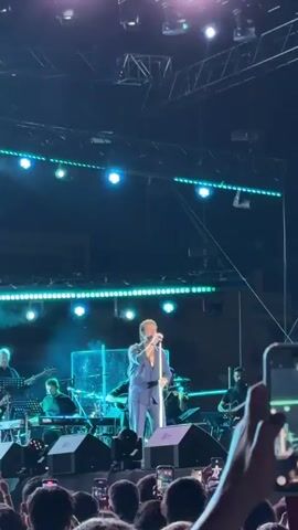 A male singer in a blue suit is performing on a stage with a band. The stage is lit with bright lights, and there is a crowd of people in the foreground, many of whom are holding up their phones to record the performance. The singer is holding a microphone and appears to be singing. The band members are playing various instruments, including guitars, keyboards, and drums. The overall atmosphere seems to be a concert or live music event.