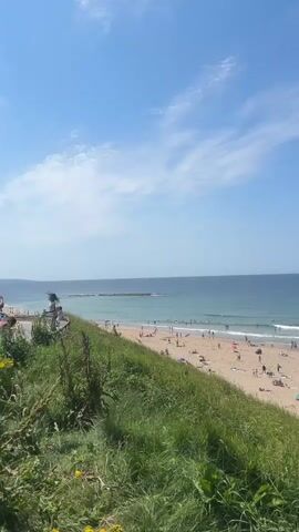 This is a vertical, wide-angle, outdoor shot of a crowded beach on a sunny day. The camera is positioned on a grassy hillside looking down at the beach and the ocean. The sky is a bright blue with scattered white clouds. The ocean is a deep blue with white waves breaking on the shore. The beach is filled with people sunbathing, sitting in chairs, and swimming in the ocean. There are umbrellas and towels on the sand. The grassy hillside in the foreground is tall and green.