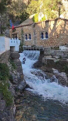 The video shows a small waterfall cascading over a rocky stream, with a stone building situated behind it. The building has a flag atop it and a few windows with blue shutters. The architecture features a mix of natural stone and some white-painted accents. The surrounding area is characterized by lush greenery. A person on a motorcycle is riding through the middle of the shot, but is partly obscured behind the water. The sky is not clearly visible, but the lighting suggests it's a bright day.