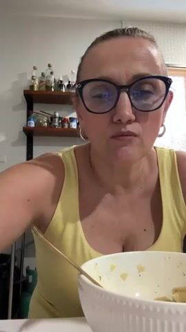 A woman with blonde hair pulled back, wearing blue glasses and a yellow tank top, is eating pasta from a white bowl with a gold spoon. She looks directly at the camera as she takes a bite. Behind her, on the wall, are two shelves holding bottles and jars of various condiments.