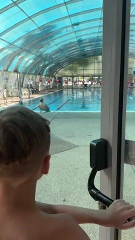 The video shows an indoor swimming pool with a blue translucent curved roof. Sunlight streams through the roof, illuminating the water. The pool has lanes marked with red and white buoys. In the water, a few people are swimming. One person is near the left side of the pool, and another person is further down the pool. Several people are sitting or standing around the edges of the pool, and there are chairs and other pool equipment visible. The foreground of the video is partially obscured by what appears to be the back of a child's head. The overall scene depicts a typical public indoor swimming pool environment.