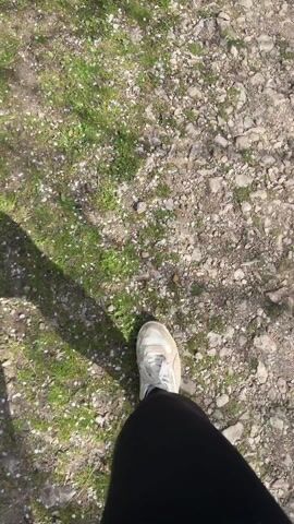 The video shows a person walking along a dirt and grass path in a wooded area. The camera is positioned from a first-person point of view, looking down at the path and then tilting upwards to show the surroundings. The path is lined with trees and bushes, some of which have white flowers in bloom. The sky is visible through the trees, with some clouds. The person is wearing black pants and white sneakers. The path appears to lead towards a distant hill or mountain.