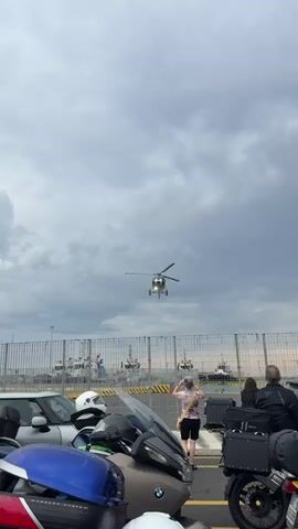 The video shows a helicopter hovering in the air, viewed from a parking area near a fence, potentially a port or similar location, as evidenced by the ships visible in the background. Several people are observing the helicopter, with one person in a pink floral shirt taking photos or videos with a mobile phone. Several motorcycles and a car are parked in the foreground. The sky is overcast with grey clouds. The helicopter is white and silver.