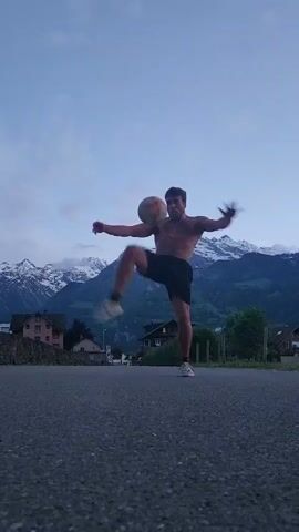 A shirtless man in black shorts juggles a soccer ball in an outdoor setting. He is positioned in the middle of a narrow asphalt road, with his arms outstretched for balance. In the background, there are buildings and trees, and snow-capped mountains are visible in the distance under a cloudy sky. After a few juggling moves, he kicks the ball away. The camera then pans down to the asphalt road.