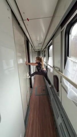 A young girl with blonde hair, wearing a dark patterned top and dark leggings, performs a split in the narrow corridor of a train car. Her left foot, clad in a white Croc, pushes against a compartment door/wall, while her right foot, in a white sneaker, rests on the handrail alongside the windows. She uses both hands to steady herself against the wall.

The train interior features a wooden-patterned floor, white walls, and large windows with white curtains. A metal handrail runs along the window side, with vents visible underneath. Compartment doors line the opposite side of the corridor. The scene is brightly lit, suggesting daytime. The girl maintains a focused expression during her stretch. A partially obscured red sign with the number "169" is visible on the lower left wall. The train appears to be in motion, indicated by ambient sounds. The setting is within a passenger train carriage.