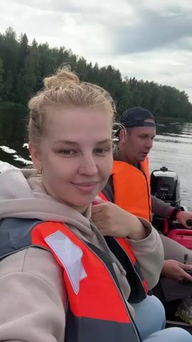 A video opens with a selfie-style shot of a young woman with blonde hair pulled back in a bun, smiling and sticking her tongue out. She is wearing a beige hooded sweatshirt and an orange and gray life jacket. Behind her, a man with short brown hair and a dark baseball cap is visible, also wearing an orange life jacket. He is looking towards the camera with a neutral expression. They are in a small motorboat on a body of water, with a lush green forest visible in the background. The sky is overcast. The man appears to be operating the boat, with a red gas can and a fishing rod visible near him. The woman makes a few different facial expressions and gestures towards the camera. The sound of the boat's motor and water can be heard, along with some indistinct chatter. The video continues with the woman and man in the boat, moving through the water.