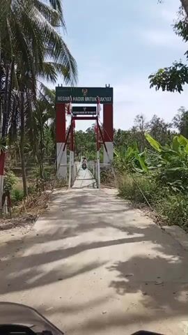 A short video captures a view from a motorcycle as it approaches a suspension bridge. The bridge is painted red and white, with a sign at the top reading "NEGAR HADIR UNTUK RAKYAT," which translates to "The State is Present for the People." Two people on motorcycles are visible on the bridge ahead. The environment is lush with tropical vegetation, including palm trees and banana plants. The sky is bright blue with some clouds.