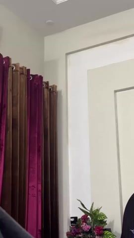 A video taken from a low angle shows a person's head and shoulders on the right side of the frame. They are wearing a dark top. The person is partially obscured by a decorative wall panel. In the background, a set of curtains hangs on the left. The curtains are a deep purple on the left side and a dark brown on the right. A small table with a plant and some flowers is visible in the lower right corner of the frame. The lighting in the room appears to be from overhead.