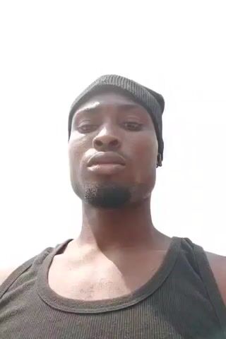 A low-angle shot shows a dark-skinned man wearing a black beanie and a black tank top. He looks up at the sky, then speaks, gesturing with his right hand. He then puts his hand to his chin. The sky is bright and mostly clear with some scattered clouds.