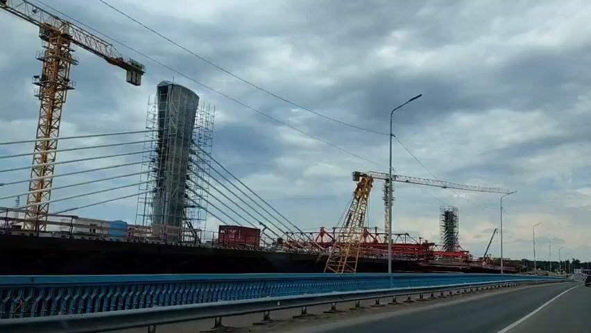 This video shows a view of a large construction project, likely a bridge, from a moving vehicle. The scene is dominated by a massive cable-stayed bridge under construction. Several large cranes, including a yellow tower crane and a longer, horizontal crane, are visible, along with scaffolding and other construction equipment. The bridge deck appears to be in progress, with sections of structural steel and concrete visible. The sky is overcast with dramatic clouds. The road in the foreground has a blue railing, suggesting the vantage point is from another elevated structure or roadway. The overall impression is one of significant engineering activity and progress.